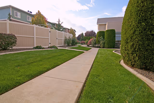 A paved walkway winding through a well-maintained outdoor garden area with green grass, trimmed bushes, and a beige fence on the left side. Residential-style buildings are visible in the background under a partly cloudy sky.