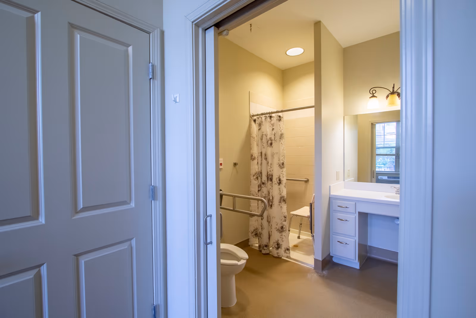 View into a bathroom with a toilet, a shower area with a floral shower curtain and a shower chair, a vanity with drawers and a large mirror, and a window reflecting outside light. The bathroom has beige walls and a brown floor.