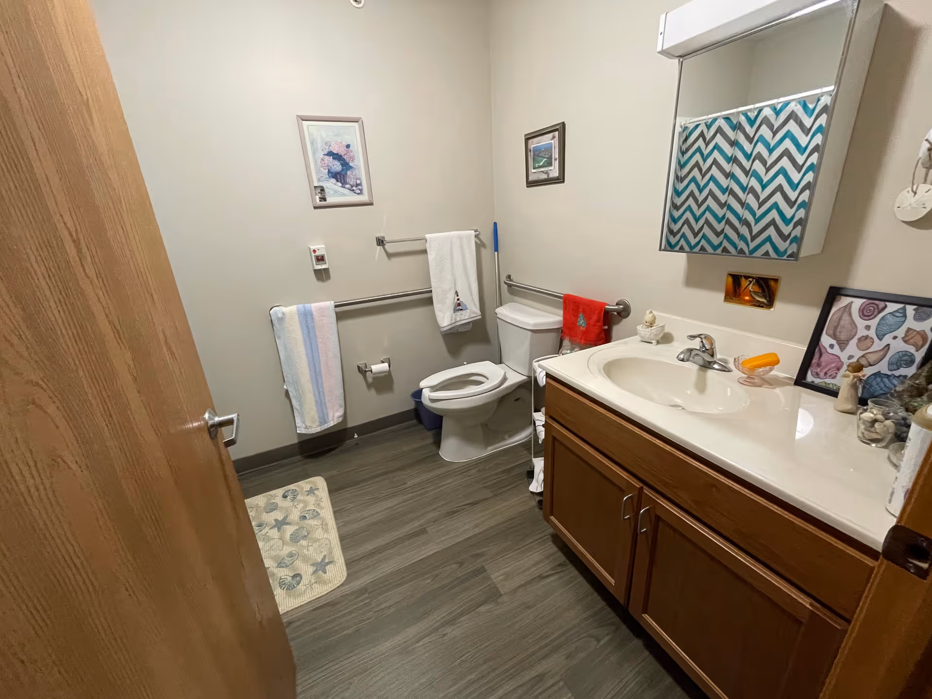 A small bathroom with a wooden door partially open, a toilet with grab bars on the wall, a sink with a wooden cabinet underneath, a mirror above the sink with a chevron patterned shower curtain reflected in it, and various decorative items including framed pictures and towels.