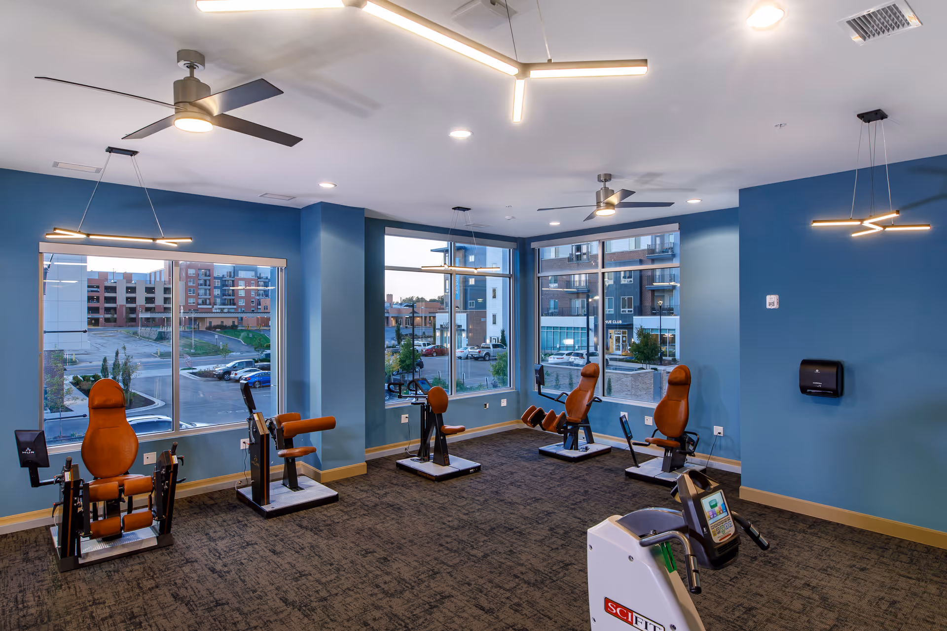 A fitness room with several exercise machines designed for seated workouts, featuring brown padded seats and black frames. The room has large windows showing an outside view of a parking lot and nearby buildings. The walls are painted blue, and the ceiling has modern light fixtures and ceiling fans.