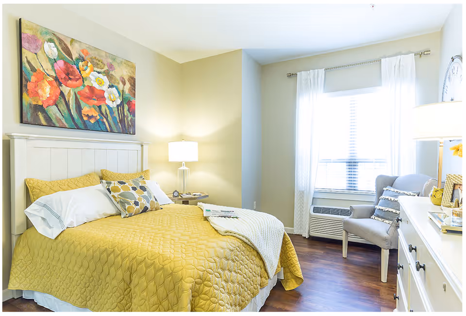 A bright and cozy bedroom featuring a white wooden bed frame with a yellow quilted bedspread and decorative pillows. A colorful floral painting hangs above the bed. There is a bedside table with a lamp and an alarm clock. A window with white curtains allows natural light to fill the room. A gray upholstered chair with a decorative pillow sits near the window, and a white dresser with a lamp and framed photos is visible on the right side.