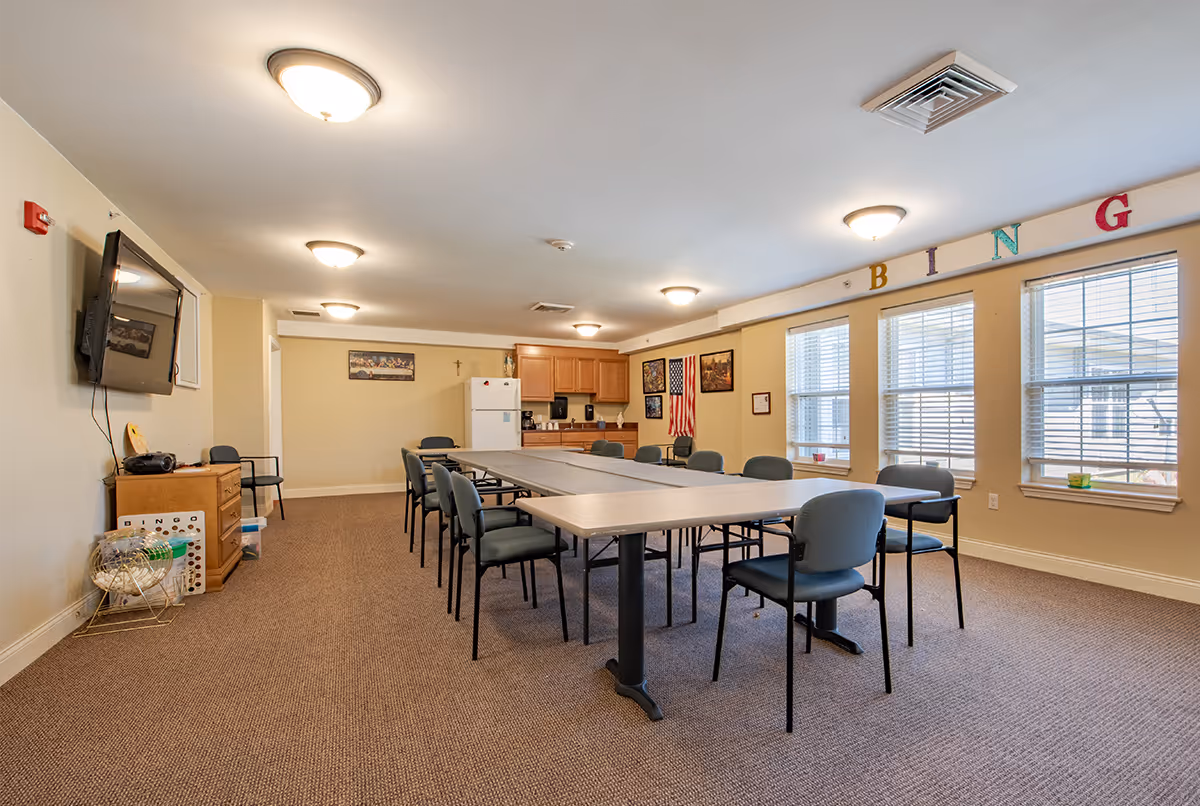 A senior living community activity room with long tables and chairs, windows along the right wall, and a kitchenette at the back with 'BINGO' letters above the windows.