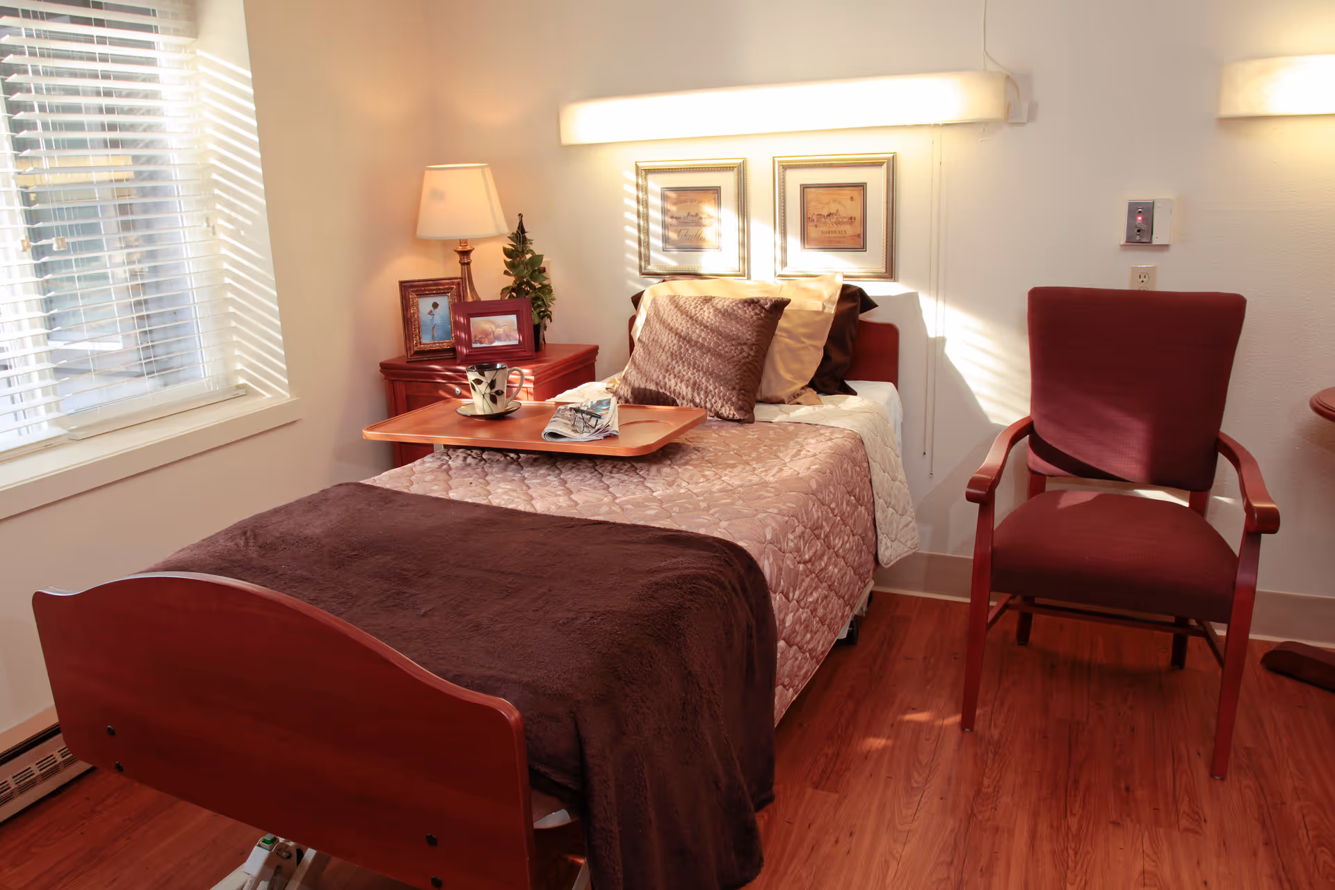A cozy bedroom in a senior living facility with a single bed covered in a brown blanket and beige quilt. There is a wooden bedside table with a lamp, framed photos, and a small plant. A wooden tray with a coffee cup and a newspaper rests on the bed. Next to the bed is a maroon upholstered chair, and the room has wooden flooring and a window with white blinds letting in natural light.