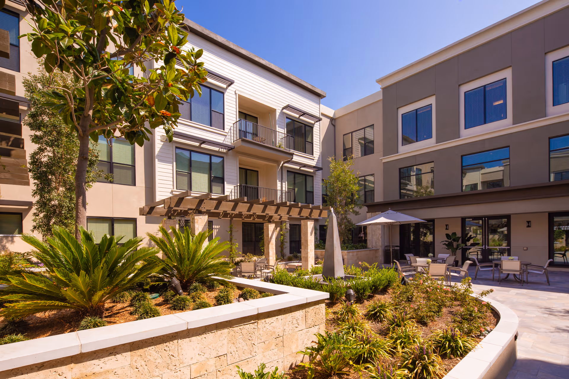 Outdoor courtyard area at Oakmont of Orange featuring landscaped garden beds with various plants and shrubs, a pergola with seating underneath, patio tables with umbrellas, and a modern three-story building with large windows surrounding the courtyard under a clear blue sky.