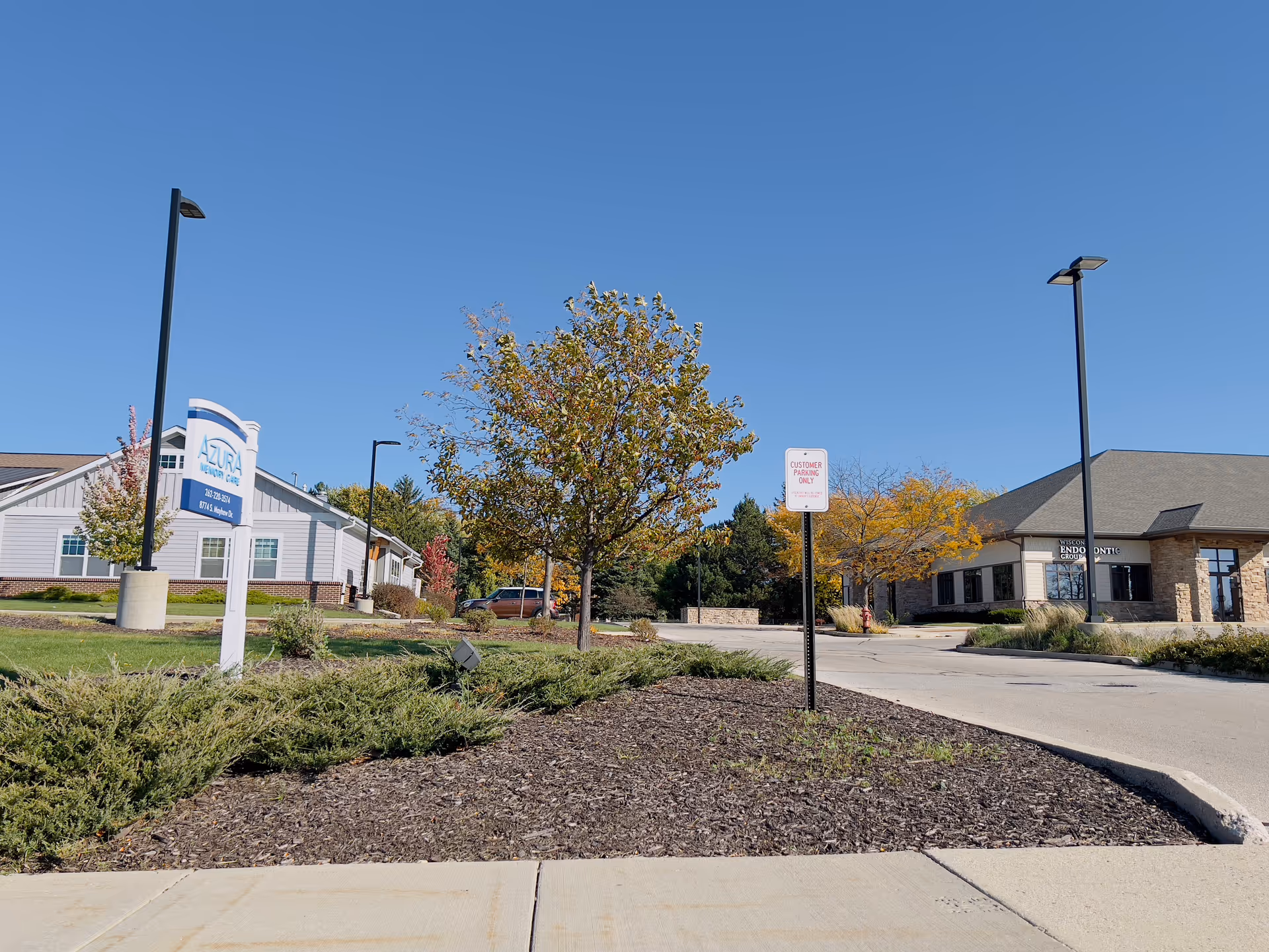 Exterior view of Azura Memory Care Of Oak Creek II facility on a clear sunny day, showing the building, a sign with the facility name and contact information, a tree with autumn leaves, and a parking area with a Customer Parking Only sign.
