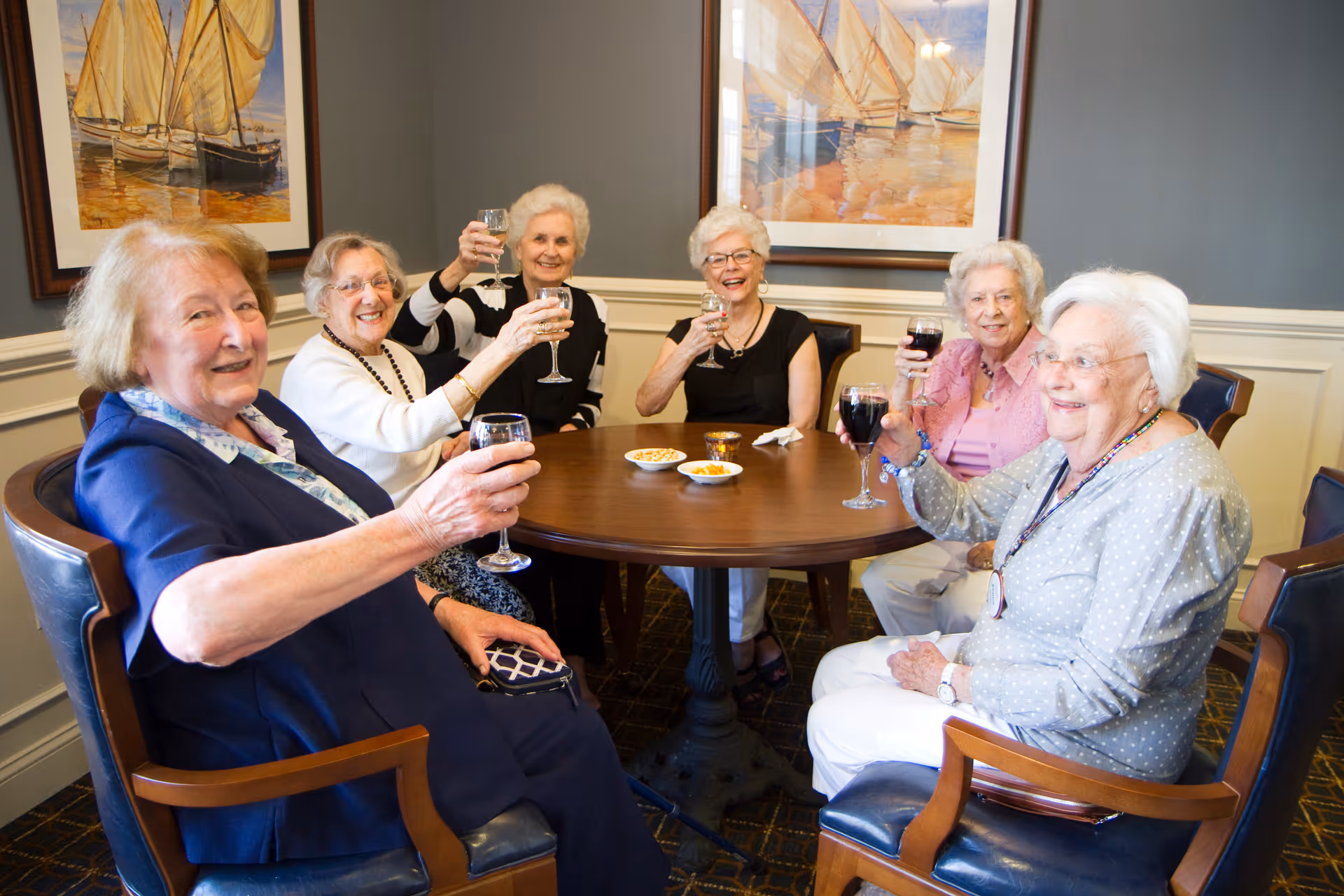 Six elderly women sitting around a round wooden table in a well-lit room, smiling and raising glasses of wine or water in a toast. The room has gray walls with white wainscoting and framed paintings of sailboats on the wall behind them. There are small bowls of snacks on the table.