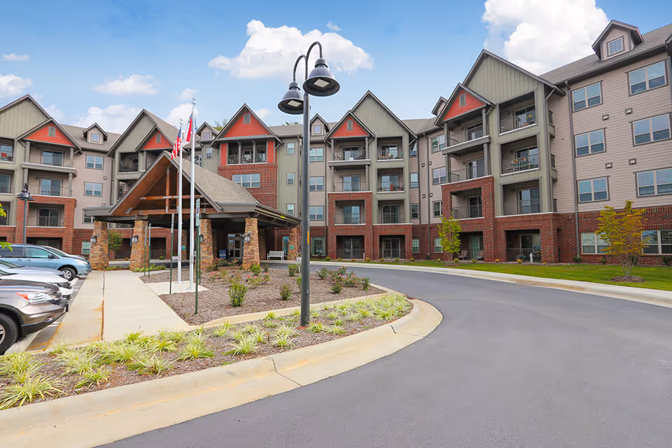 Exterior view of The Crossings At Reynolds Mountain senior living facility showing a multi-story building with balconies, a covered entrance with stone pillars, a driveway, parked cars, and landscaping under a partly cloudy sky.