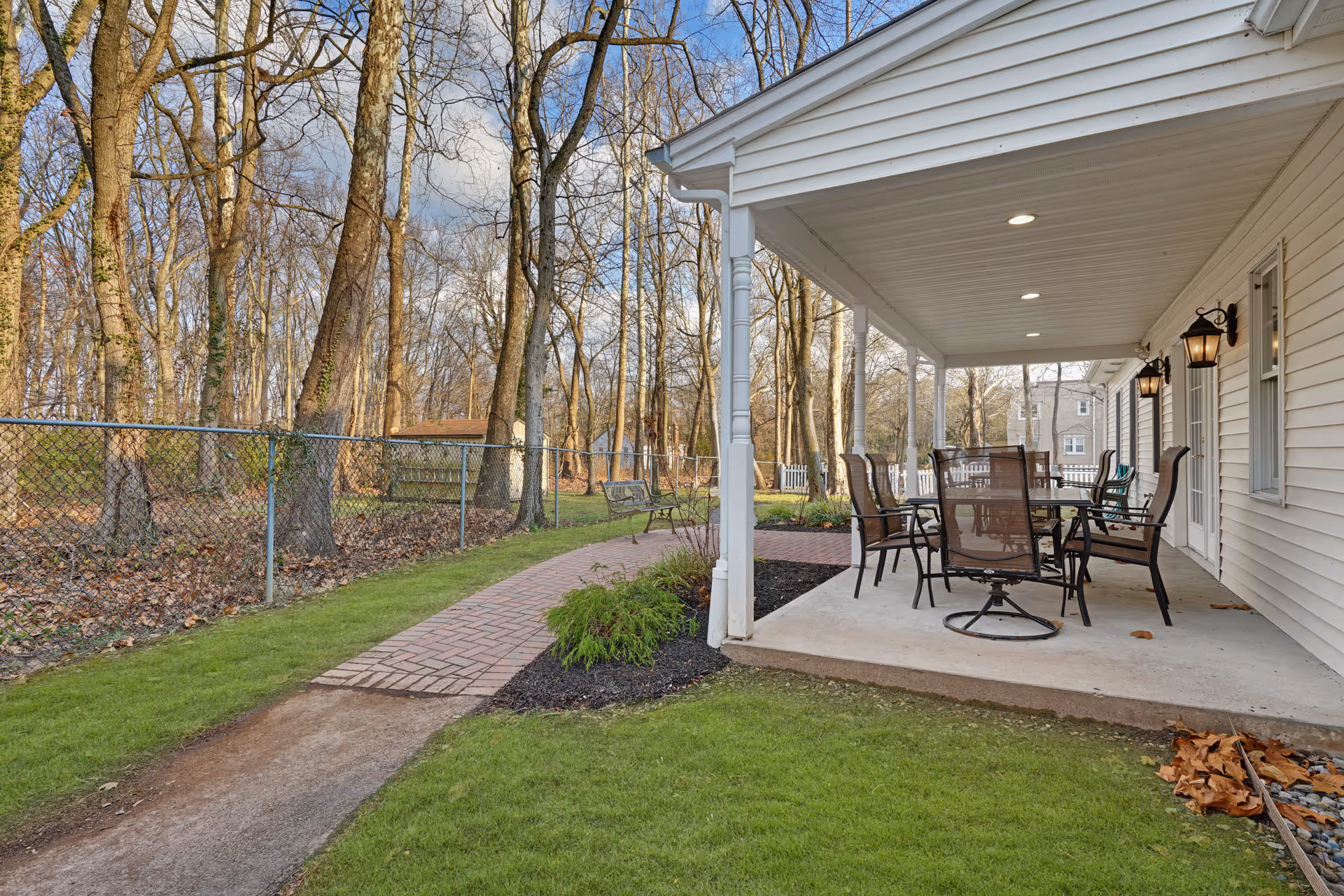 Covered outdoor patio area with a table and several chairs on a concrete floor, adjacent to a white building with wall-mounted lantern lights. A brick pathway leads away from the patio through a grassy area bordered by a chain-link fence and trees without leaves.