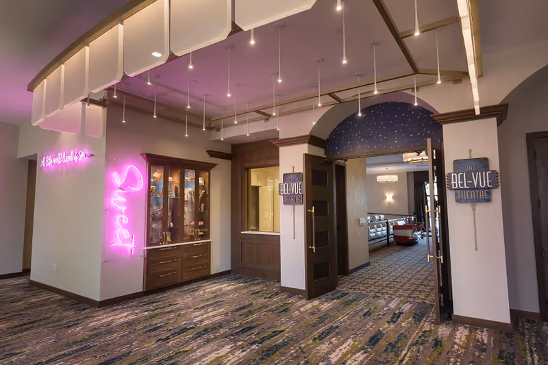 Interior view of a senior living facility hallway with a carpeted floor and a ceiling with hanging lights. On the left wall, there is a neon pink sign that says 'Sweet' and another neon sign with cursive text. A wooden display cabinet with glass doors is also visible. On the right, double doors lead into a room labeled 'The Bel-Vue Theatre' with a starry archway above the entrance.