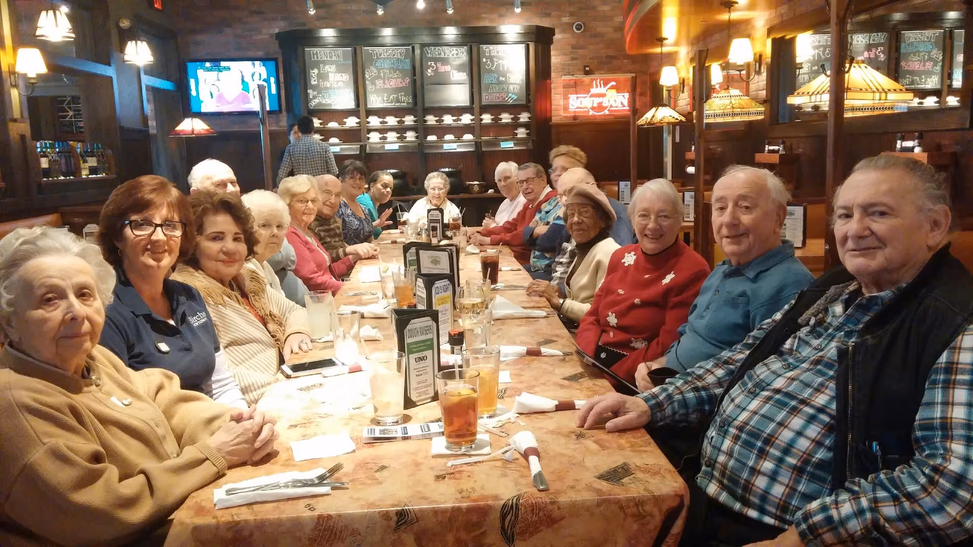 A large group of older adults seated around a long table in a warmly lit restaurant-style dining room.