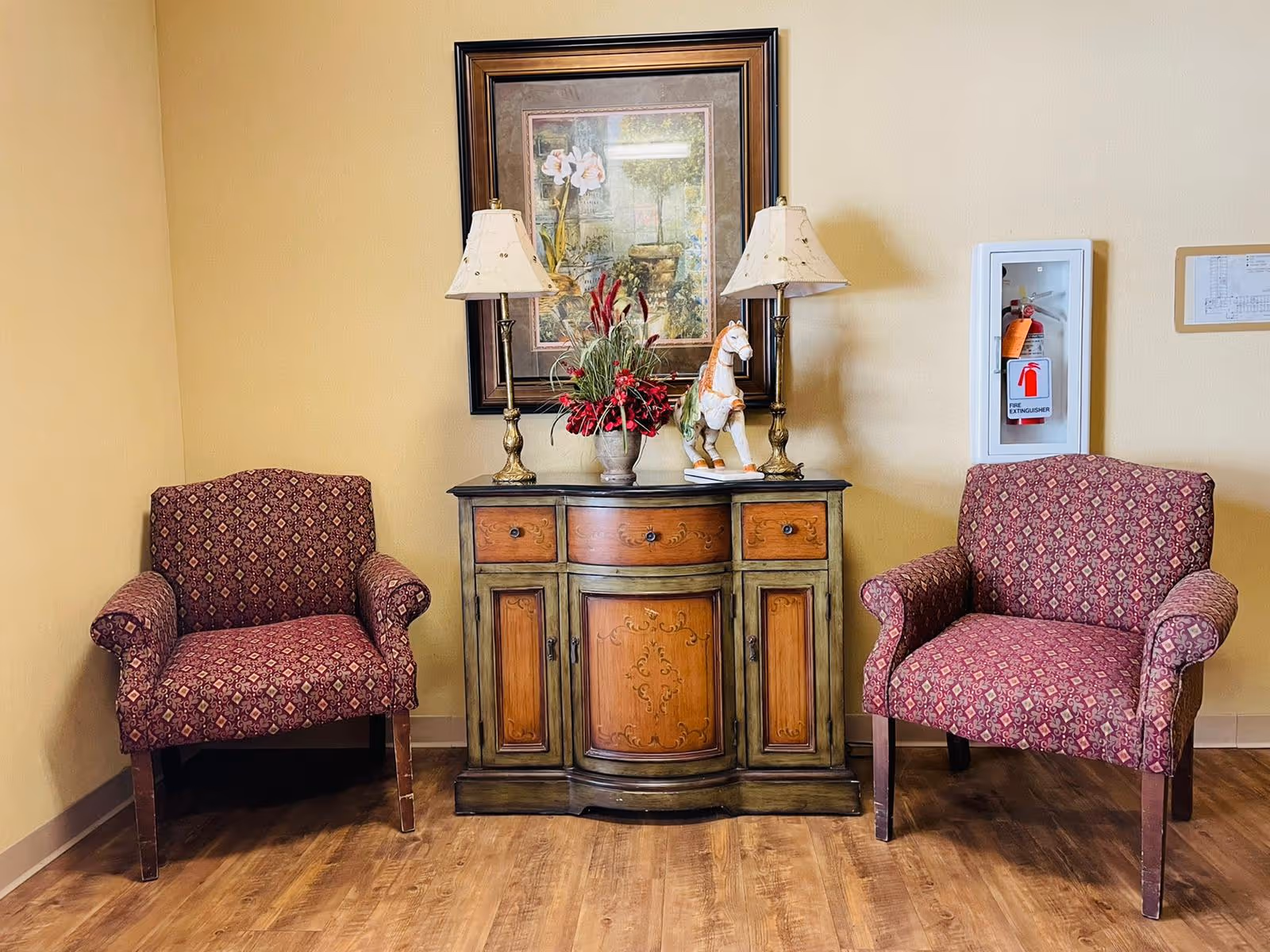 Two patterned armchairs flank a wooden sideboard with decorative carvings. On the sideboard are two table lamps, a vase with red flowers, and a white horse figurine. Above the sideboard hangs a framed floral painting. A fire extinguisher is mounted on the wall to the right.