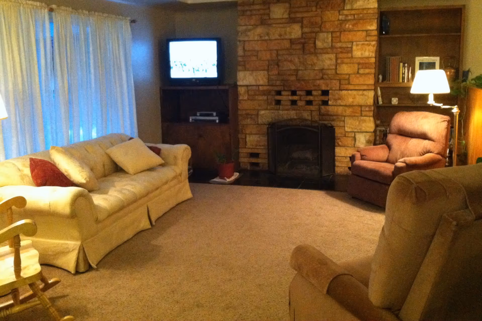 A cozy living room with a beige sofa adorned with cushions, a brown armchair, a rocking chair, a stone fireplace, a wooden bookshelf with books and a lamp, and a TV on a wooden stand near a window with white curtains.