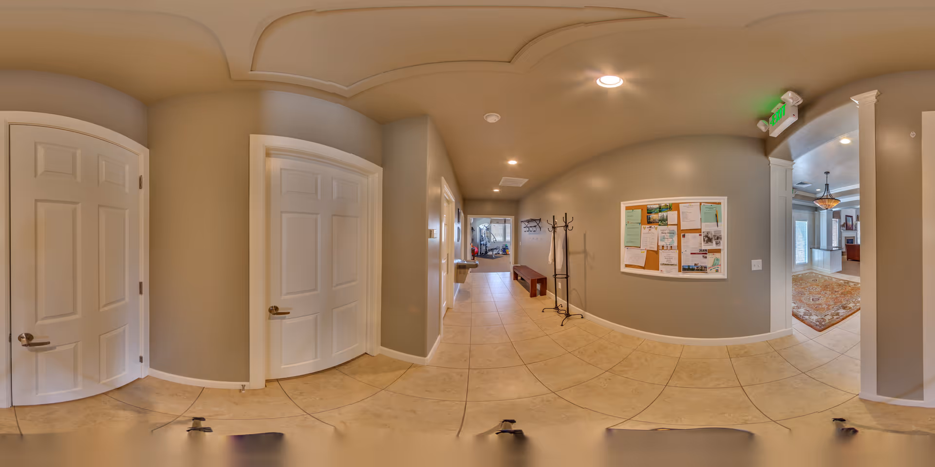 A wide hallway in a senior living facility with beige tiled floors and light gray walls. There are two white doors on the left side and a bulletin board with various notices on the right wall. A coat rack and a bench are positioned along the right side of the hallway. The hallway leads to a room with a carpet and chandelier visible in the distance.