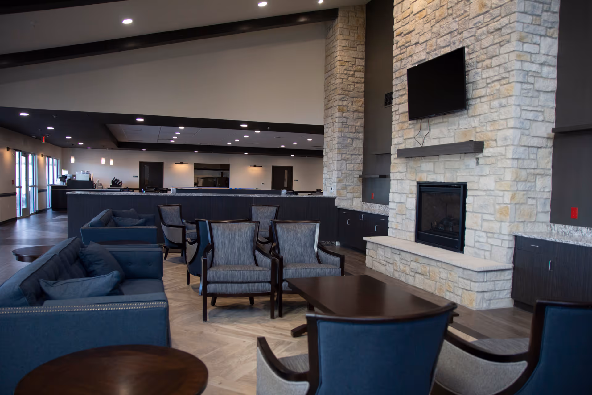 Open senior living lounge with sofas and armchairs arranged around a stone fireplace and wall-mounted TV, adjacent to a service counter.
