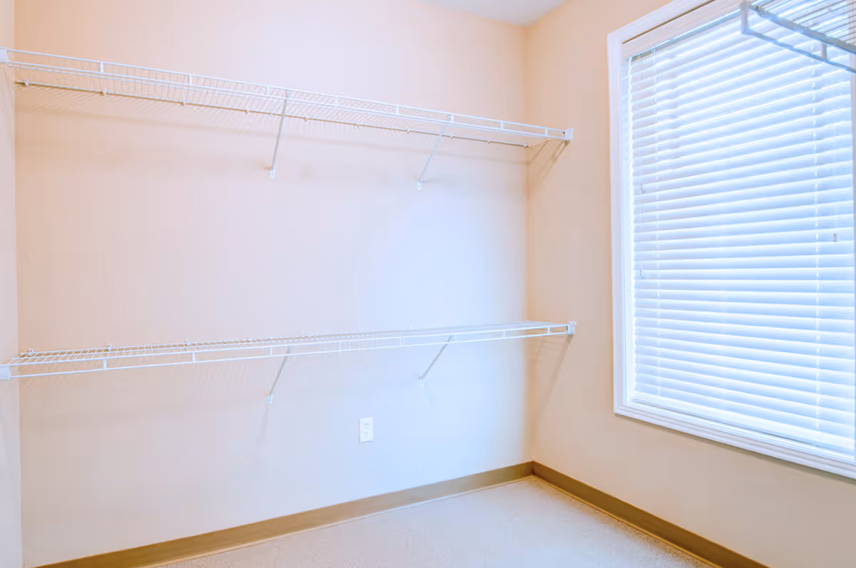 Empty room with beige walls, two white wire shelves mounted on the wall, a window with white blinds on the right side, and a beige carpeted floor.