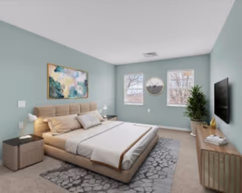 A modern bedroom with a large beige upholstered bed, two bedside tables with lamps, a patterned rug, two windows with a round mirror between them, a potted plant, a wooden dresser, and a wall-mounted flat-screen TV. The walls are painted light blue and there is a colorful abstract painting above the bed.