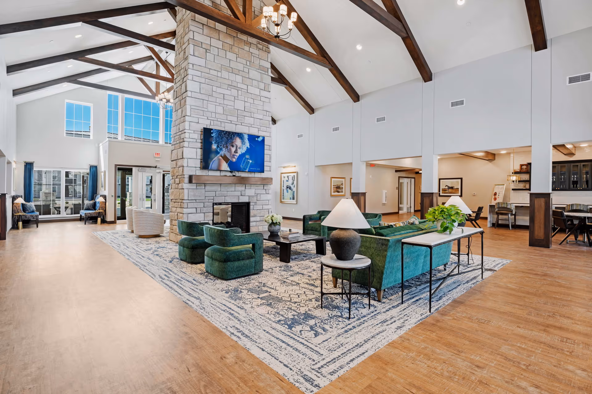 Spacious senior living facility common area with high vaulted ceilings featuring exposed wooden beams, a large stone fireplace with a mounted TV above it, green upholstered chairs and sofas arranged on a patterned area rug, side tables with lamps and plants, and large windows letting in natural light.