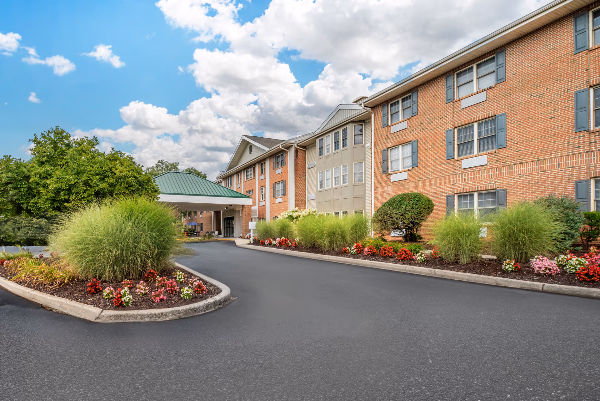 Entrance drive and landscaped front of a multi-story brick senior living building with a covered porte-cochere under a partly cloudy sky.
