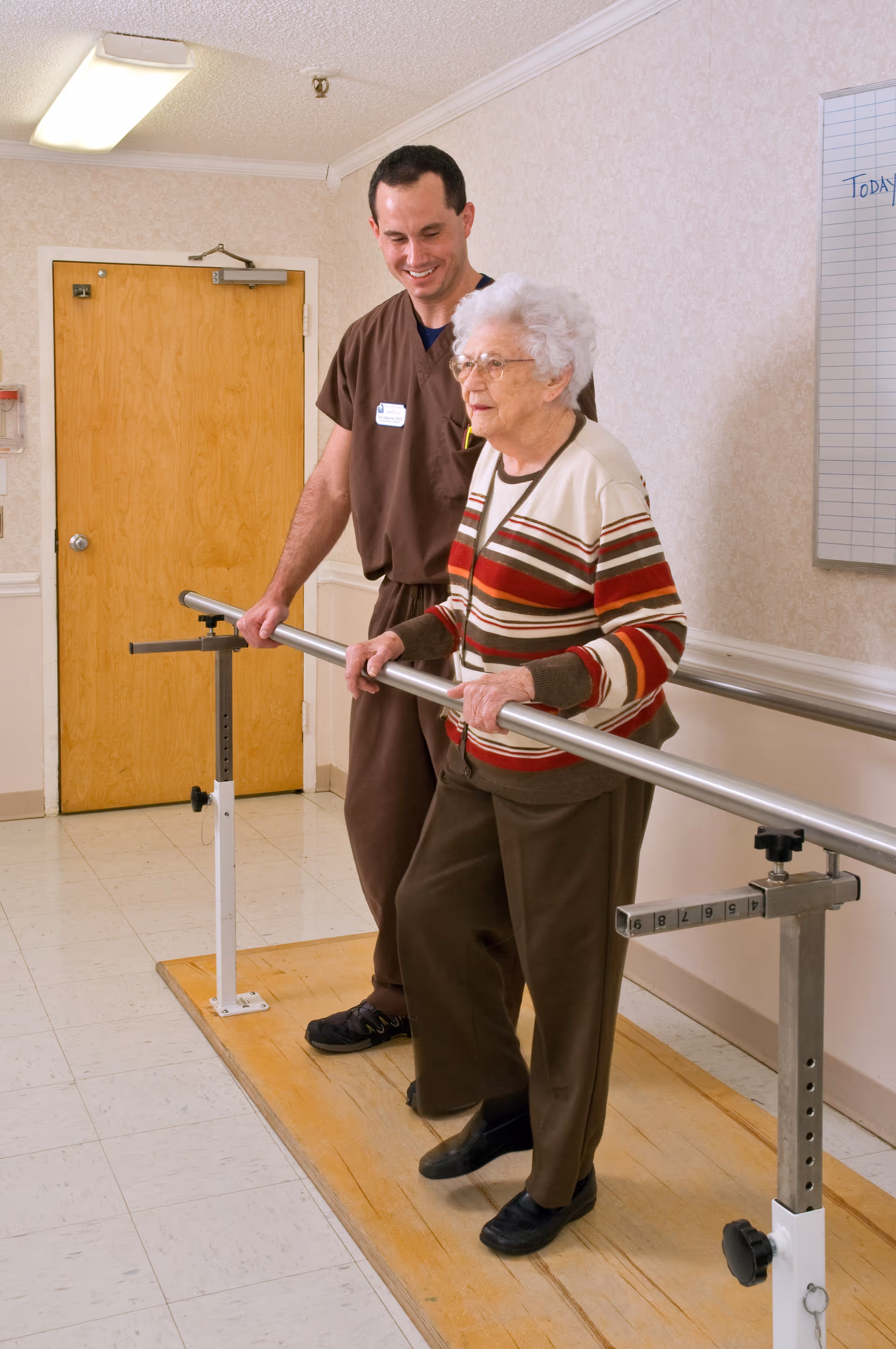 An elderly woman holding onto parallel bars while a male caregiver or physical therapist stands beside her, smiling and providing support in a rehabilitation or therapy room with a wooden door and a whiteboard on the wall.
