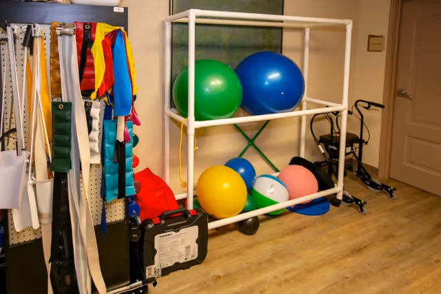 A therapy or exercise room corner with various physical therapy equipment including resistance bands hanging on a rack, colorful exercise balls on a white metal shelf, and a walker against the wall. The room has light wood flooring and beige walls.