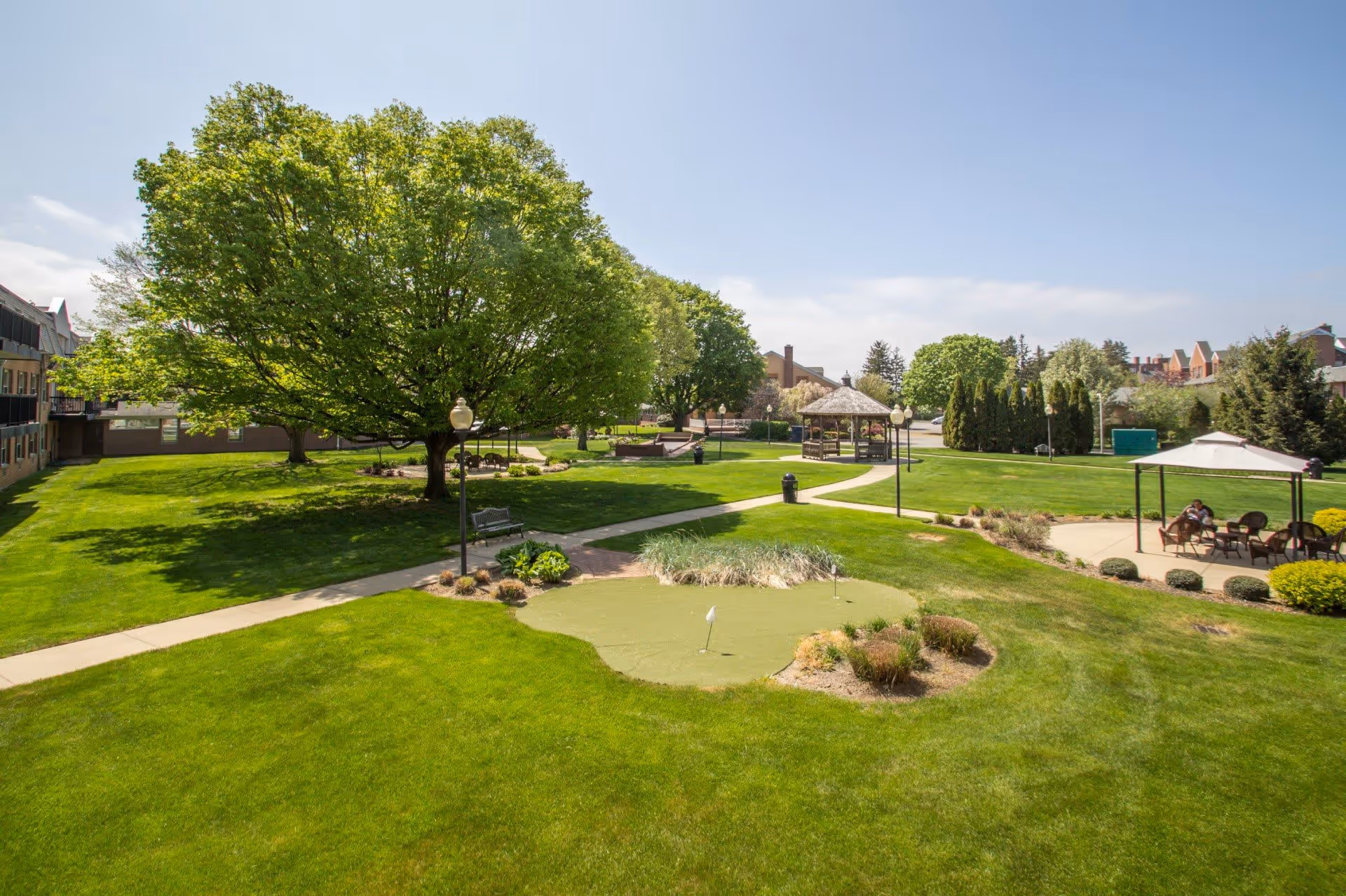 A sunny outdoor garden area at Dominican Village featuring a large green lawn with a putting green, several mature trees, paved walkways, benches, a gazebo, and a covered seating area where two people are sitting.