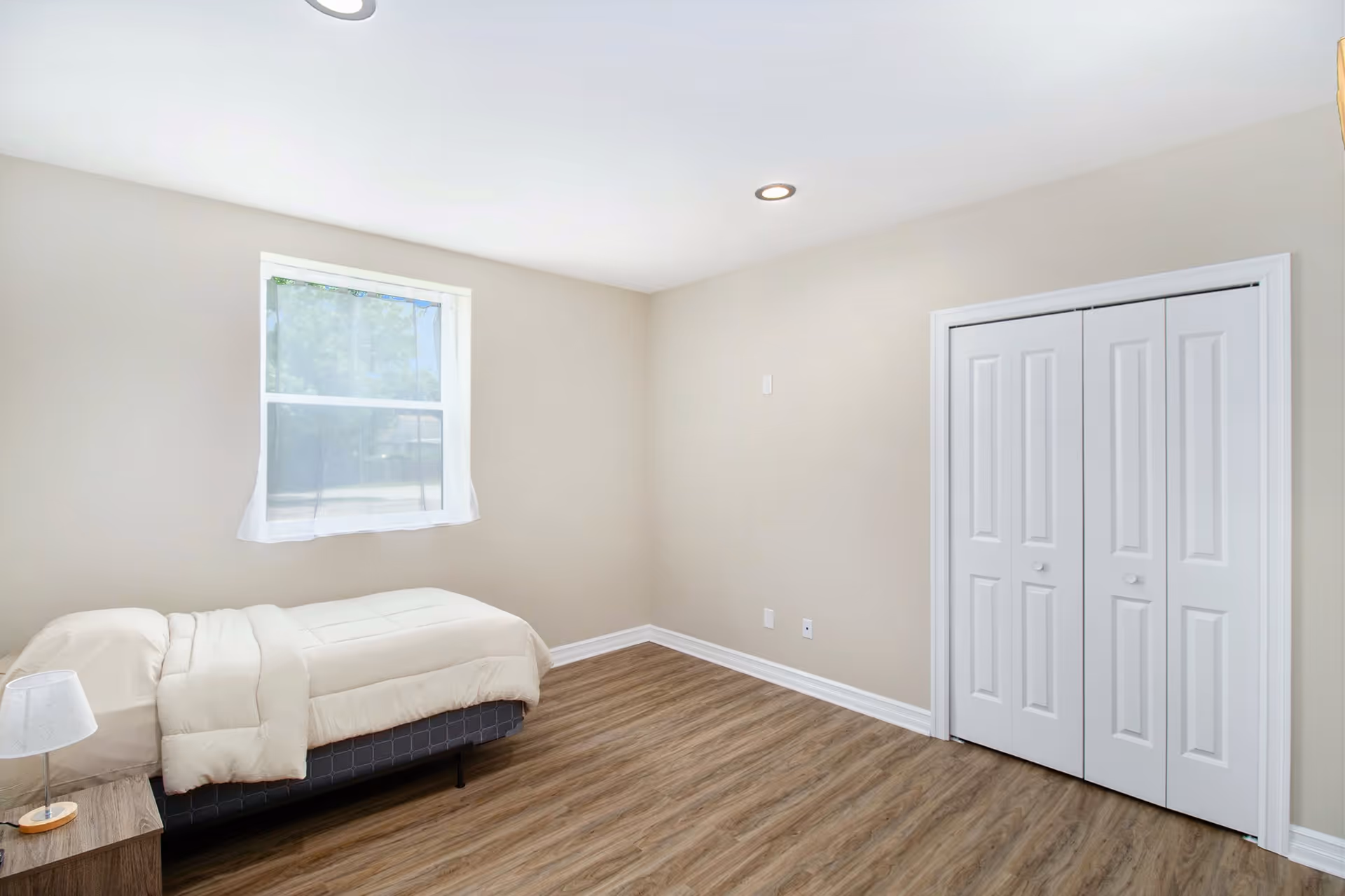 A simple bedroom with a single bed covered in beige bedding, a small wooden nightstand with a lamp, a window with sheer white curtains, beige walls, wood flooring, and a white double-door closet.