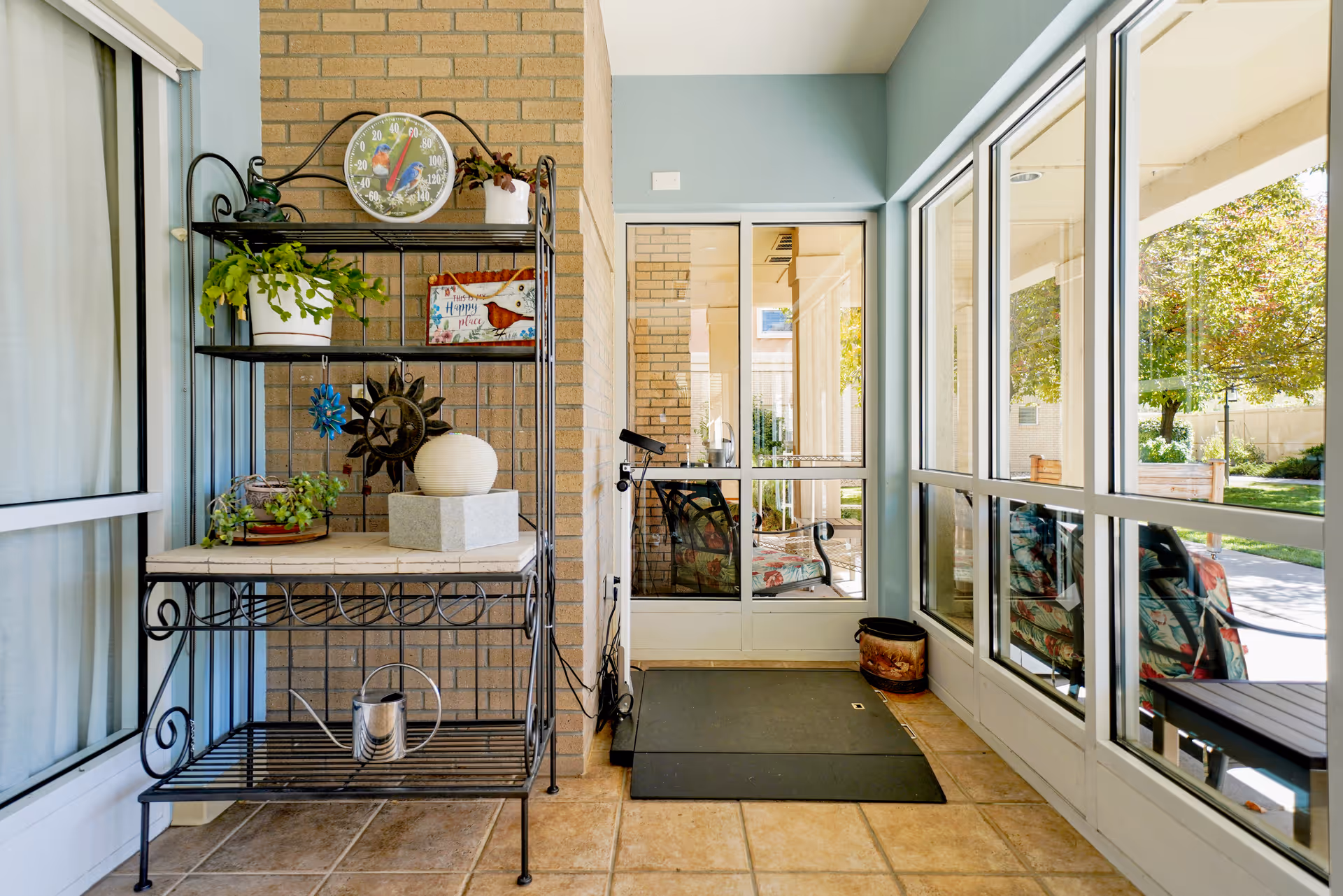 A bright indoor patio area with large windows showing an outdoor garden. There is a black metal shelving unit with plants, decorative items, and a round thermometer on the top shelf. The floor is tiled, and there is a black mat in front of a glass door leading outside.
