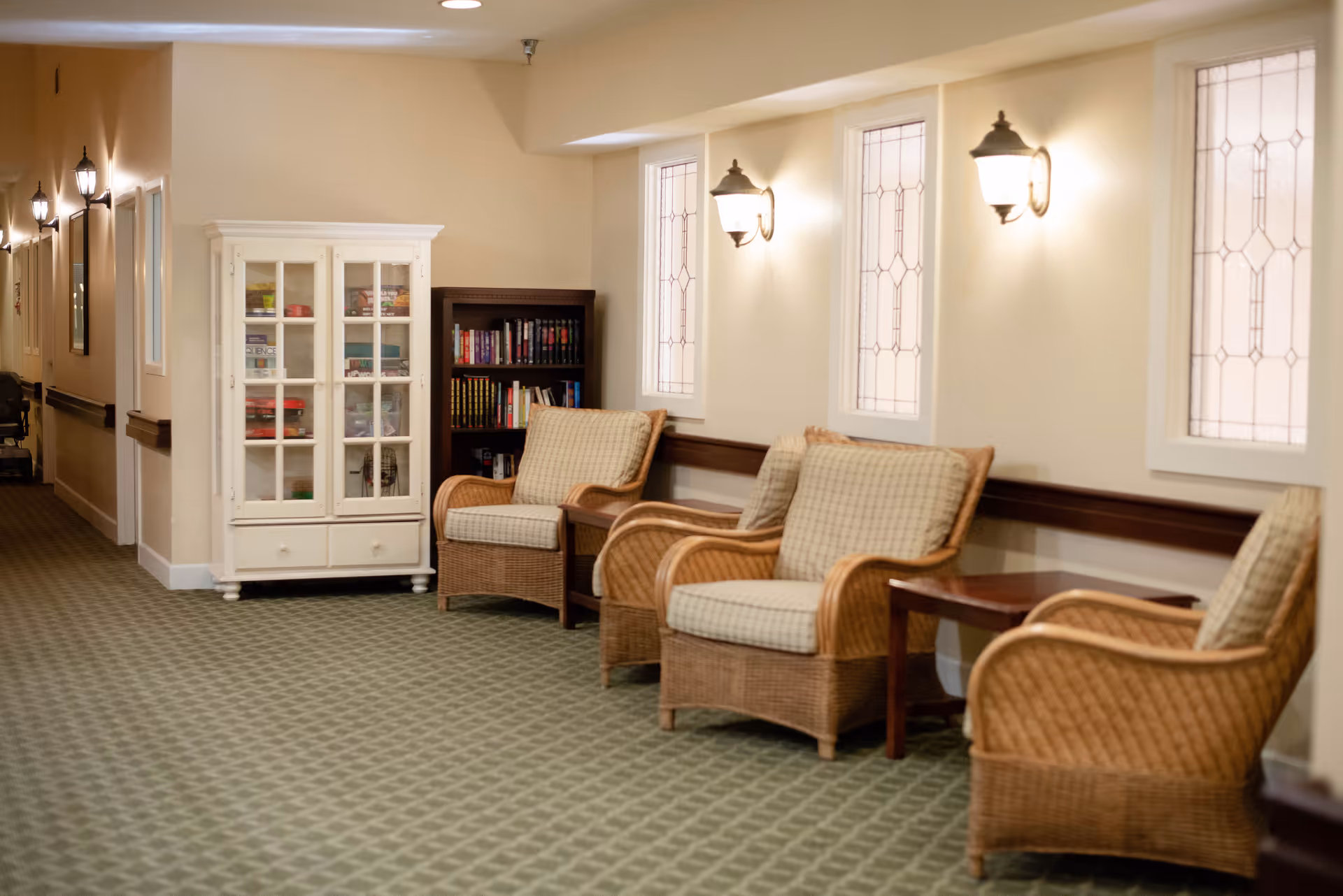 A hallway in a senior living facility with three cushioned wicker chairs arranged along the wall, small wooden side tables between them, a white cabinet with glass doors displaying various items, and a bookshelf filled with books. The walls have decorative windows and wall-mounted lamps providing warm lighting.