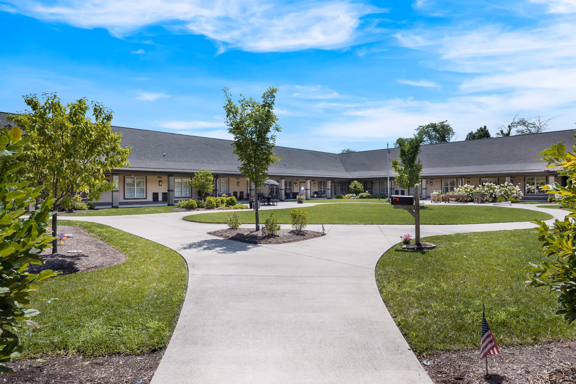 Outdoor courtyard area of Liberty Place Memory Care featuring a paved walkway that splits into two paths around a grassy lawn with small trees and shrubs. The building surrounds the courtyard with multiple windows and doors, and there are benches and a mailbox visible. The sky is clear and blue with some clouds.
