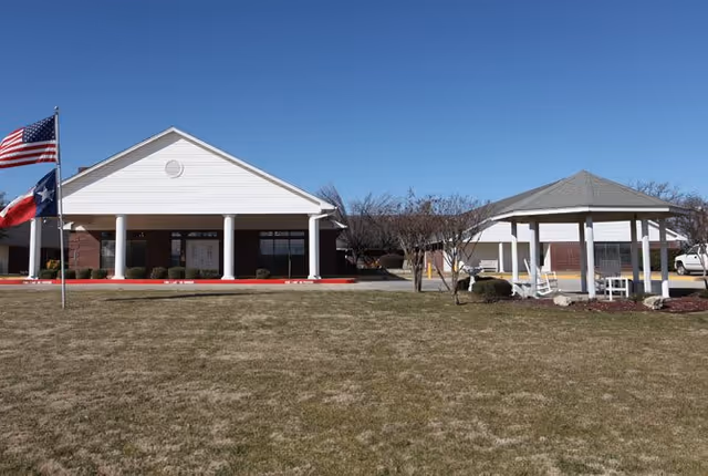 Exterior view of Lamar Place facility showing a single-story building with a white triangular roof supported by columns. There is a gazebo with rocking chairs on the right side and two flagpoles with the American and Texas flags on the left. The foreground features a large grassy area under a clear blue sky.