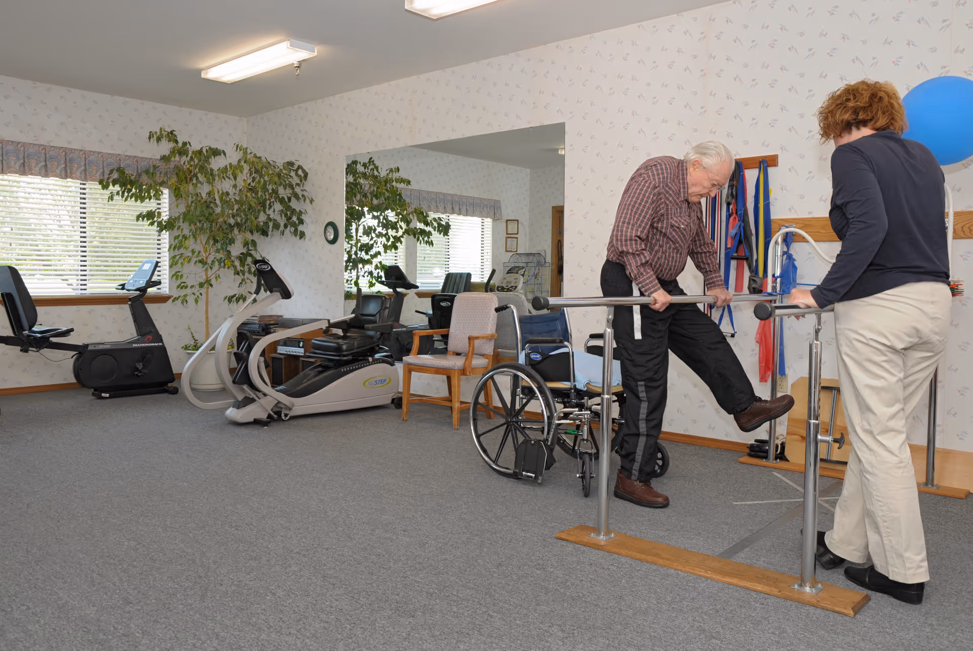 An elderly man is doing a leg exercise using parallel bars in a rehabilitation or physical therapy room, assisted by a woman. The room has exercise equipment including stationary bikes, a wheelchair, and resistance bands hanging on the wall. There is a large mirror on one wall and a window with blinds and a plant nearby.
