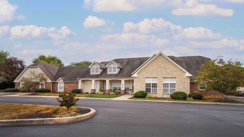 Single-story senior living facility with stone and brick facade, a covered front entrance and landscaped driveway under a partly cloudy sky.