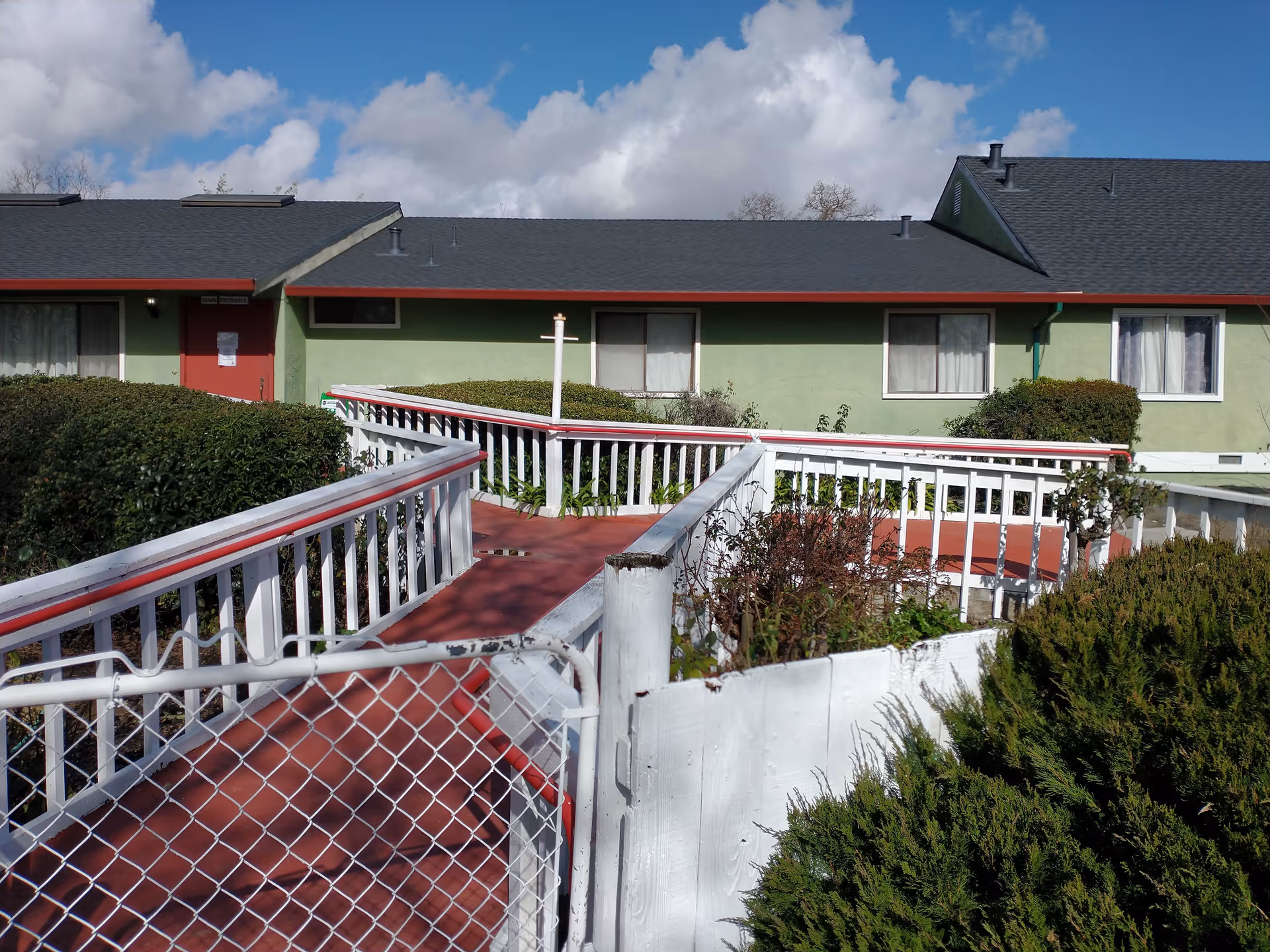 Outdoor view of a single-story building with a green exterior and dark roof under a partly cloudy blue sky. A red and white wheelchair accessible ramp with railings leads to a red door labeled 'Main Entrance'. Bushes and greenery surround the ramp and building.