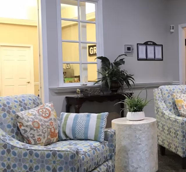 A cozy seating area in a senior living facility with two patterned armchairs featuring decorative pillows, a small round table with a potted plant, and a wooden console table with another plant. A window with multiple panes looks into another room, and a menu board is mounted on the wall.