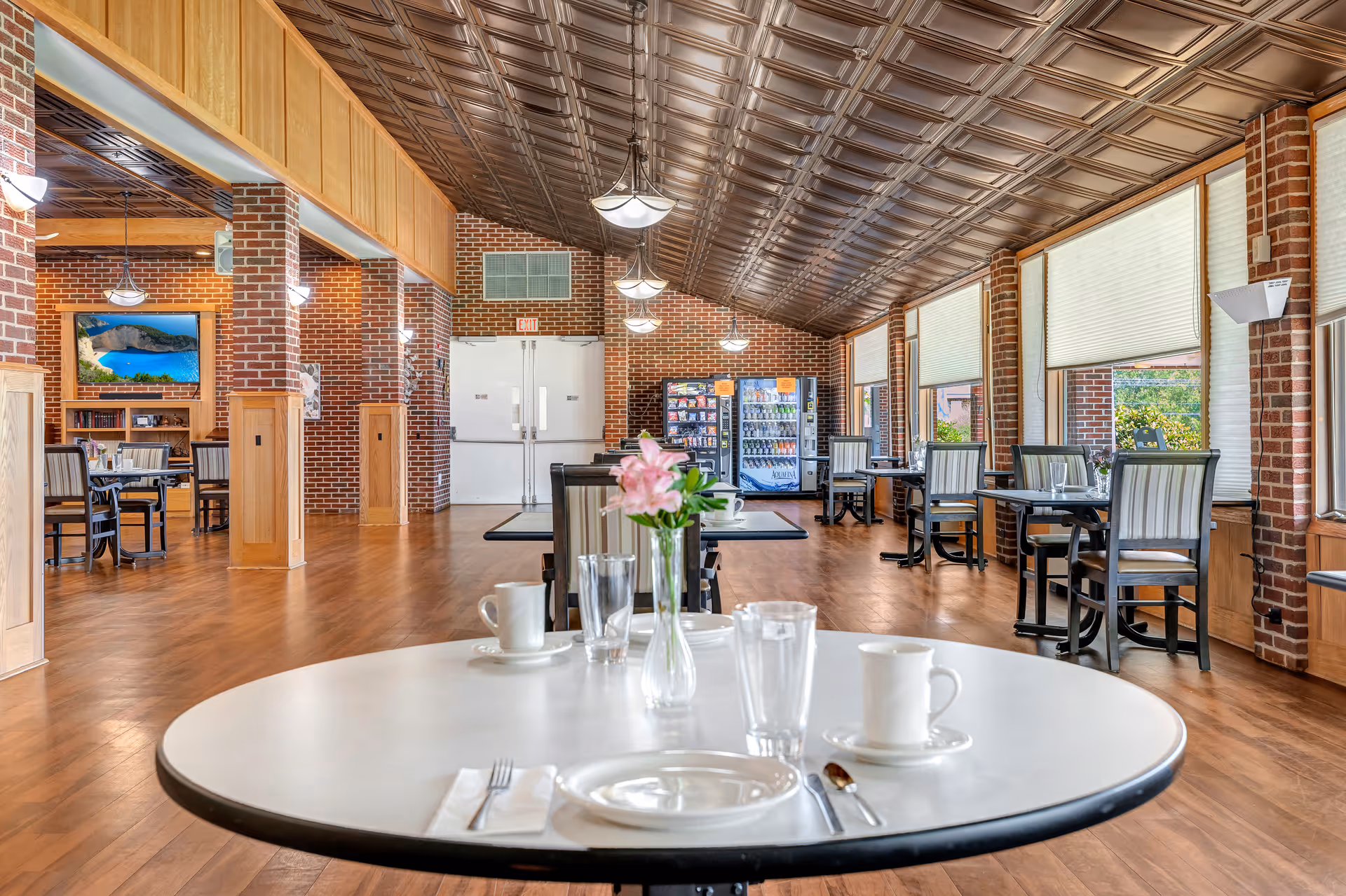 A spacious dining area with wooden floors and brick walls. Several tables and chairs are arranged throughout the room, with a round table in the foreground set with white plates, cups, glasses, and a small vase with pink flowers. Large windows with blinds line one side of the room, and vending machines are visible at the far end. The ceiling has a decorative metal pattern with hanging light fixtures.