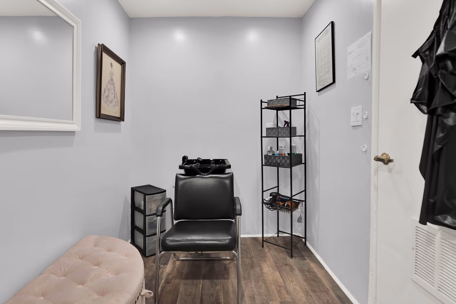 Small room with a black salon chair facing a black hair washing sink, a pink cushioned bench, a small black storage drawer unit, and a tall black metal shelving unit holding various hair care products and tools. The walls are light gray with framed artwork and a mirror, and the floor is wooden.