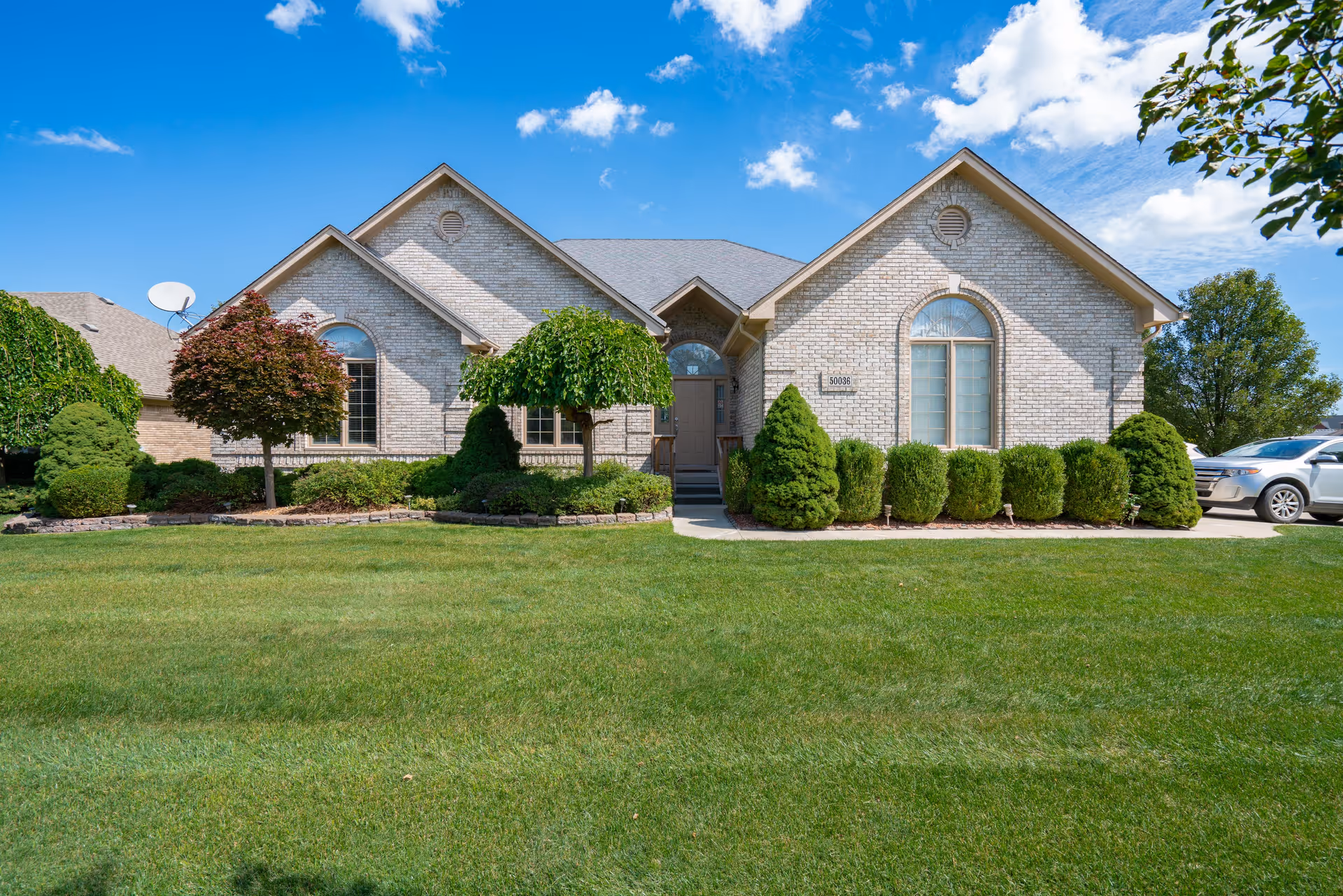 Front exterior view of a single-story brick building with a well-maintained lawn and neatly trimmed bushes and trees under a blue sky with some clouds. A driveway with a parked car is visible on the right side.