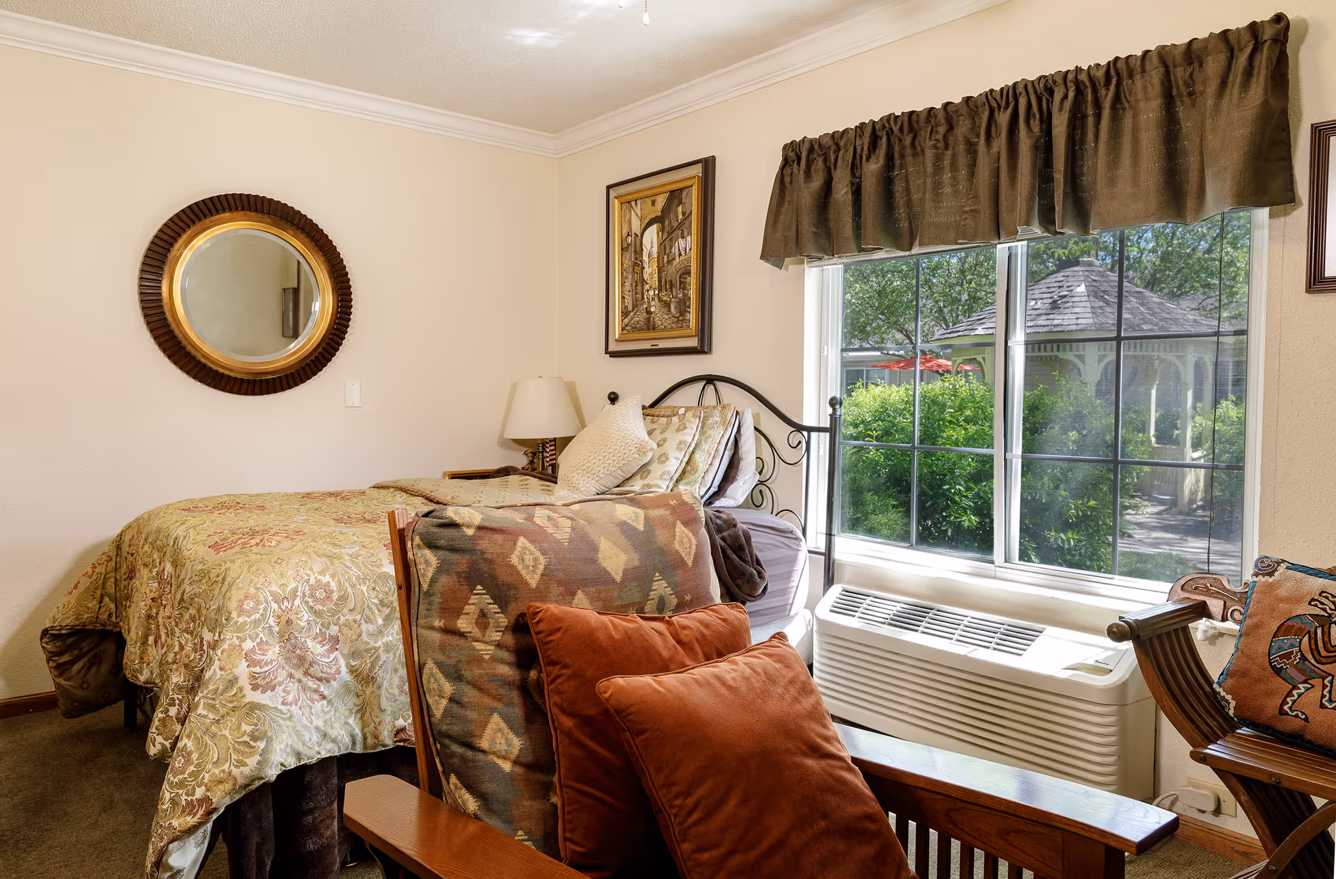 A cozy bedroom with a single bed covered in a floral patterned bedspread. There is a wooden armchair with patterned cushions and a rust-colored pillow in the foreground. A round mirror hangs on the wall above a bedside table with a lamp. A framed painting is on the wall above the bed. A large window with brown curtains shows a view of greenery and a gazebo outside. Below the window is an air conditioning unit.
