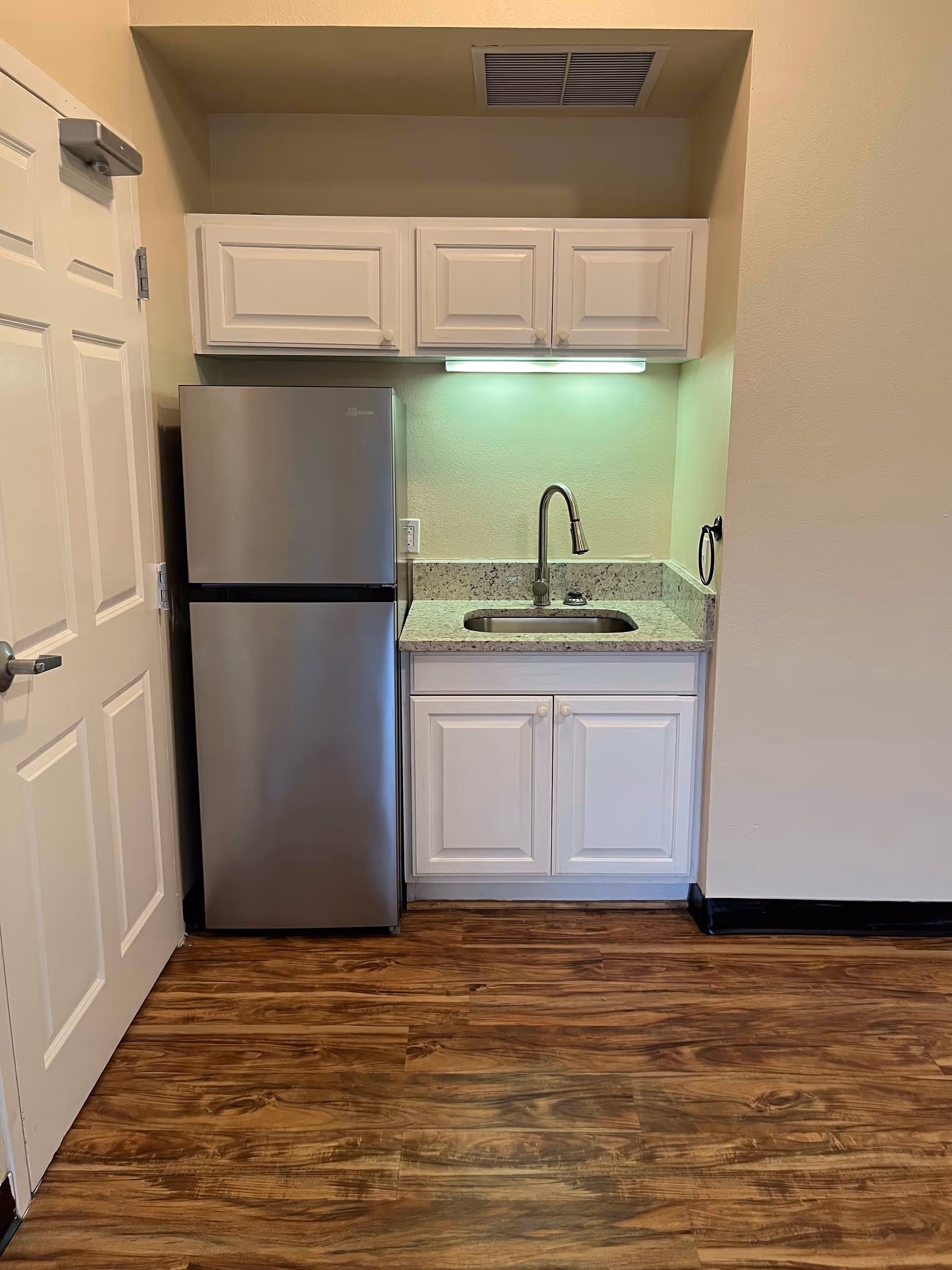 Small kitchenette area with a stainless steel refrigerator on the left, white cabinets above and below a granite countertop with a sink and a modern faucet. The floor is wood-style laminate and there is a white door partially visible on the left side.