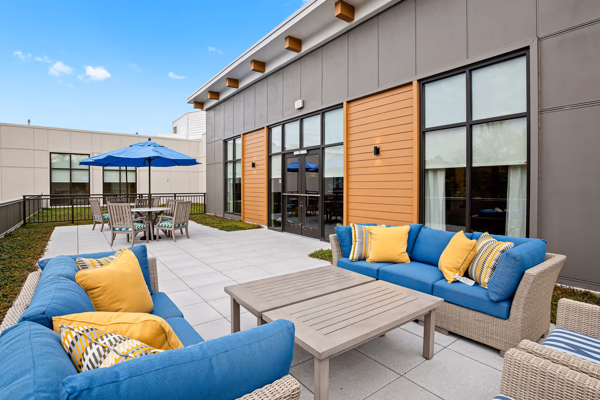 Modern outdoor patio with blue cushioned sofas, a coffee table, and a dining table with a blue umbrella outside a building with large windows.