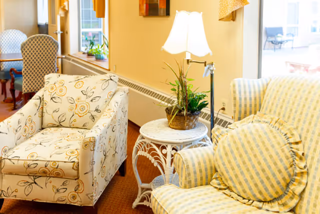 A cozy seating area in a senior living community featuring a floral patterned armchair, a yellow and blue checkered armchair with a round cushion, a white wicker side table with a potted plant and a lit table lamp, and a window in the background letting in natural light.