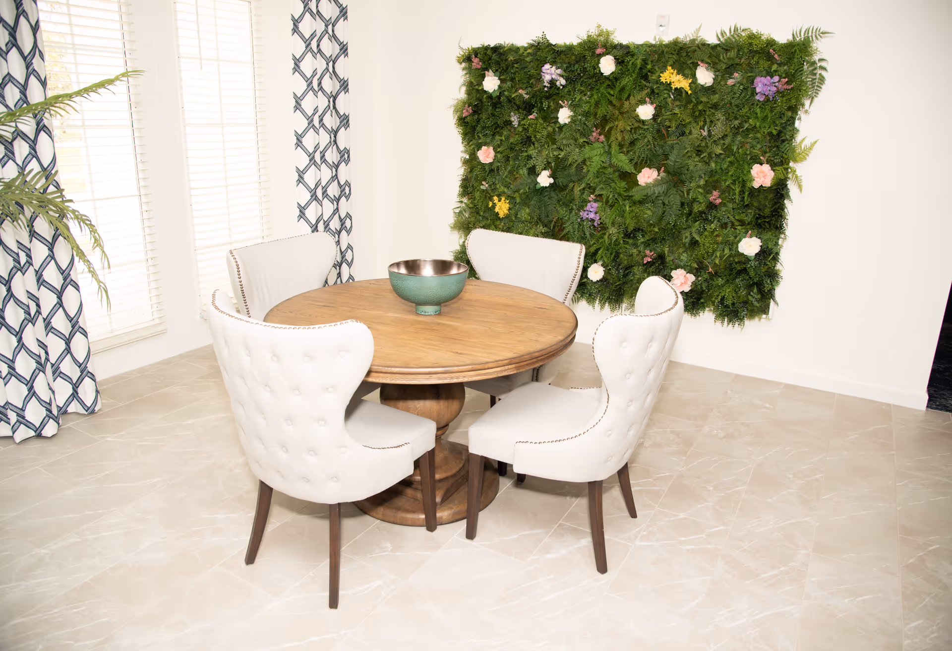 A bright dining area with a round wooden table surrounded by four upholstered cream-colored chairs. A green decorative bowl sits in the center of the table. Behind the table, a wall-mounted green plant panel with various colorful flowers adds a natural touch. Large windows with white blinds and patterned curtains allow natural light to fill the room. The floor is covered with light-colored tiles.