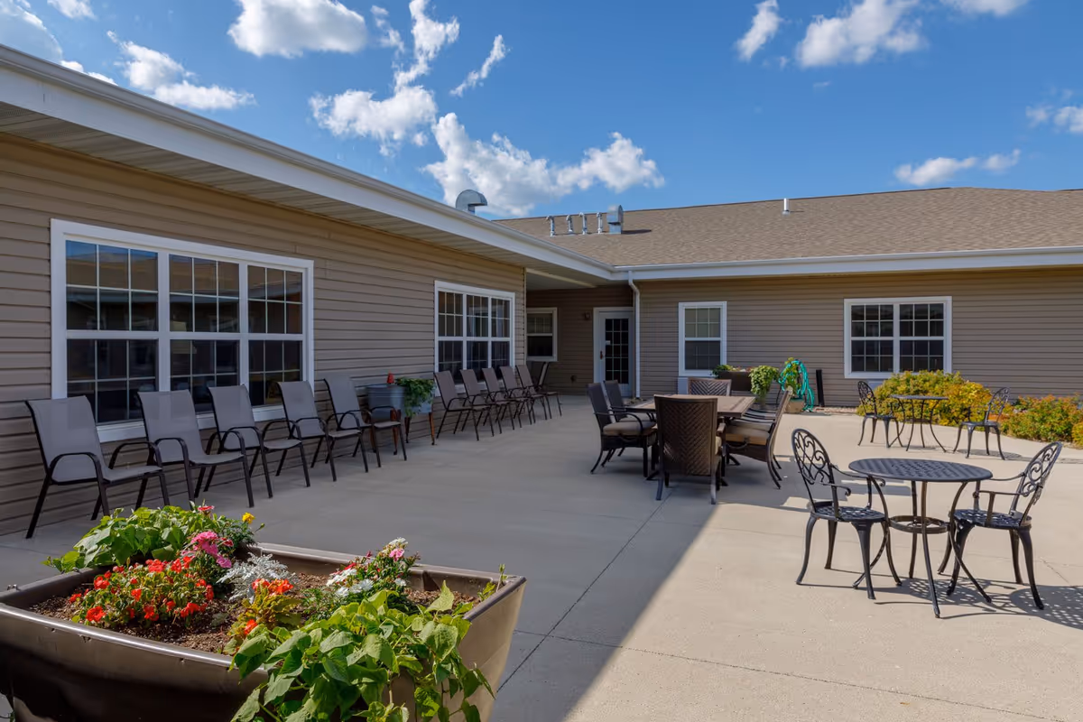 Outdoor patio area at Homestead Assisted Living & Memory Care of Algona with multiple seating arrangements including rows of chairs along the building wall, round metal tables with chairs, and a larger table with cushioned chairs. There are flower planters with colorful flowers in the foreground and a clear blue sky with some clouds above.