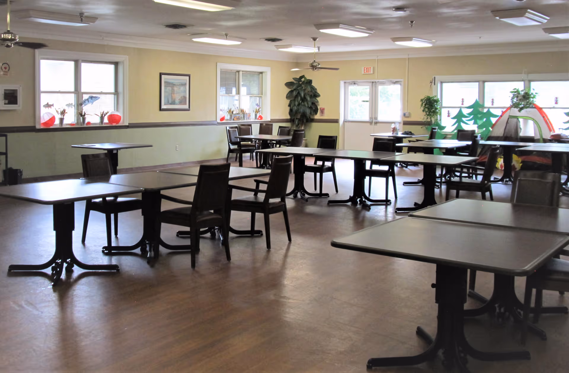 A spacious dining room with multiple tables and chairs arranged neatly. The room has large windows with decorations on the sills, a potted plant near the exit door, and a small play tent with tree cutouts in one corner. The floor is wooden, and the ceiling has several light fixtures and ceiling fans.