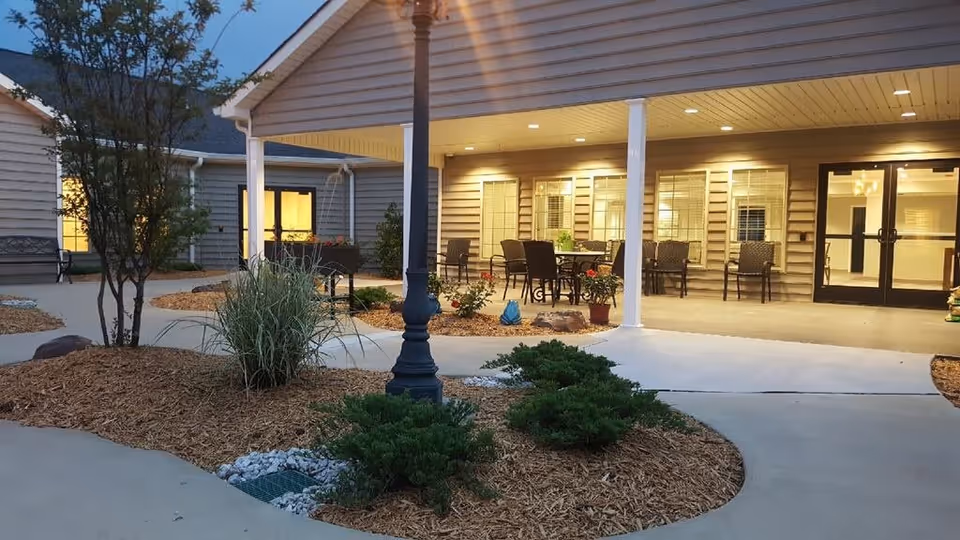 Outdoor patio area at dusk with seating arrangements including tables and chairs under a covered porch attached to a building. The area is landscaped with mulch, bushes, small trees, and decorative rocks. Warm lights illuminate the porch and windows of the building.