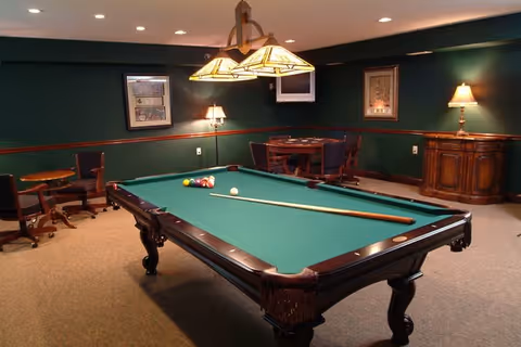 A cozy game room with a green felt pool table in the center, a cue stick resting on it, and a rack of billiard balls arranged. The room has dark green walls with wood paneling, framed artwork, a small round table with chairs, a wooden cabinet, and warm lighting from ceiling fixtures and lamps.