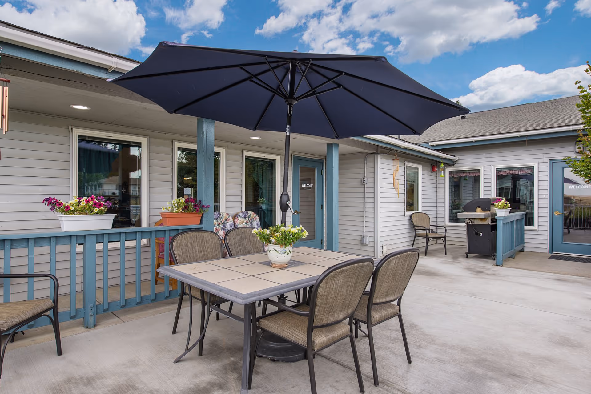 Outdoor patio area at Vista Manor with a table and six chairs under a large black umbrella. The patio is surrounded by a light gray building with blue trim, flower pots with colorful flowers on the railing, and a barbecue grill in the corner. The sky is partly cloudy.