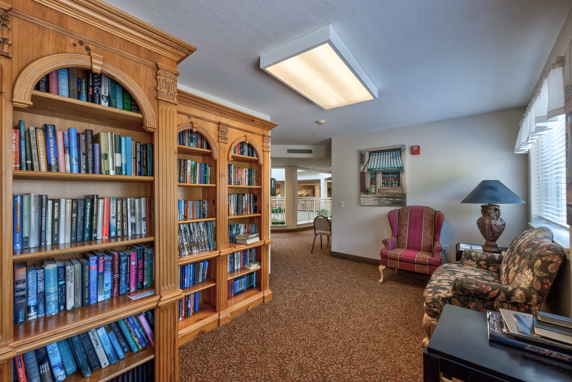 Interior view of a cozy reading area in a senior living facility featuring a large wooden bookshelf filled with books, a floral patterned sofa, a red striped armchair, a side table with a decorative lamp, and a window with white curtains allowing natural light in.