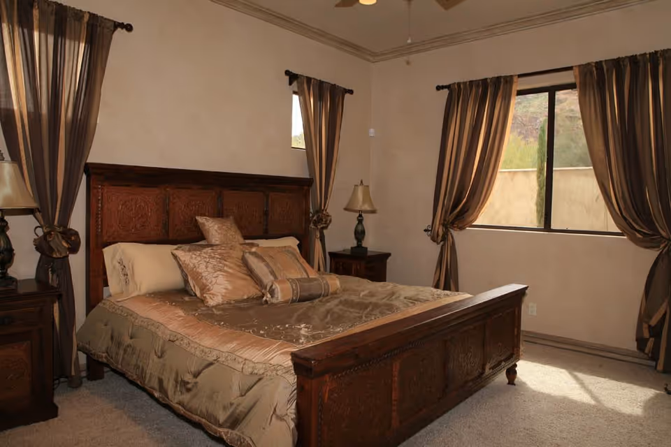 A bedroom with a large wooden bed featuring intricate carvings, dressed with beige and gold bedding and multiple decorative pillows. The room has beige walls, carpeted floor, two windows with brown curtains tied back, and matching wooden nightstands with lamps on either side of the bed.