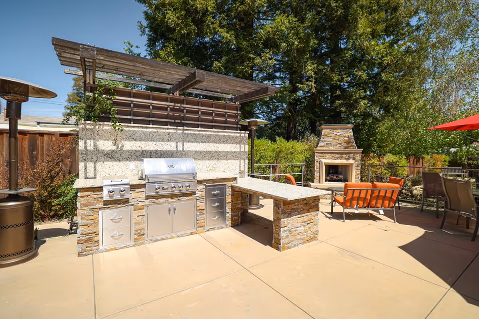 Outdoor patio area with a built-in stone and stainless steel barbecue grill, a stone countertop, patio heaters, orange cushioned seating, a stone fireplace, and surrounding trees and greenery under a clear blue sky.